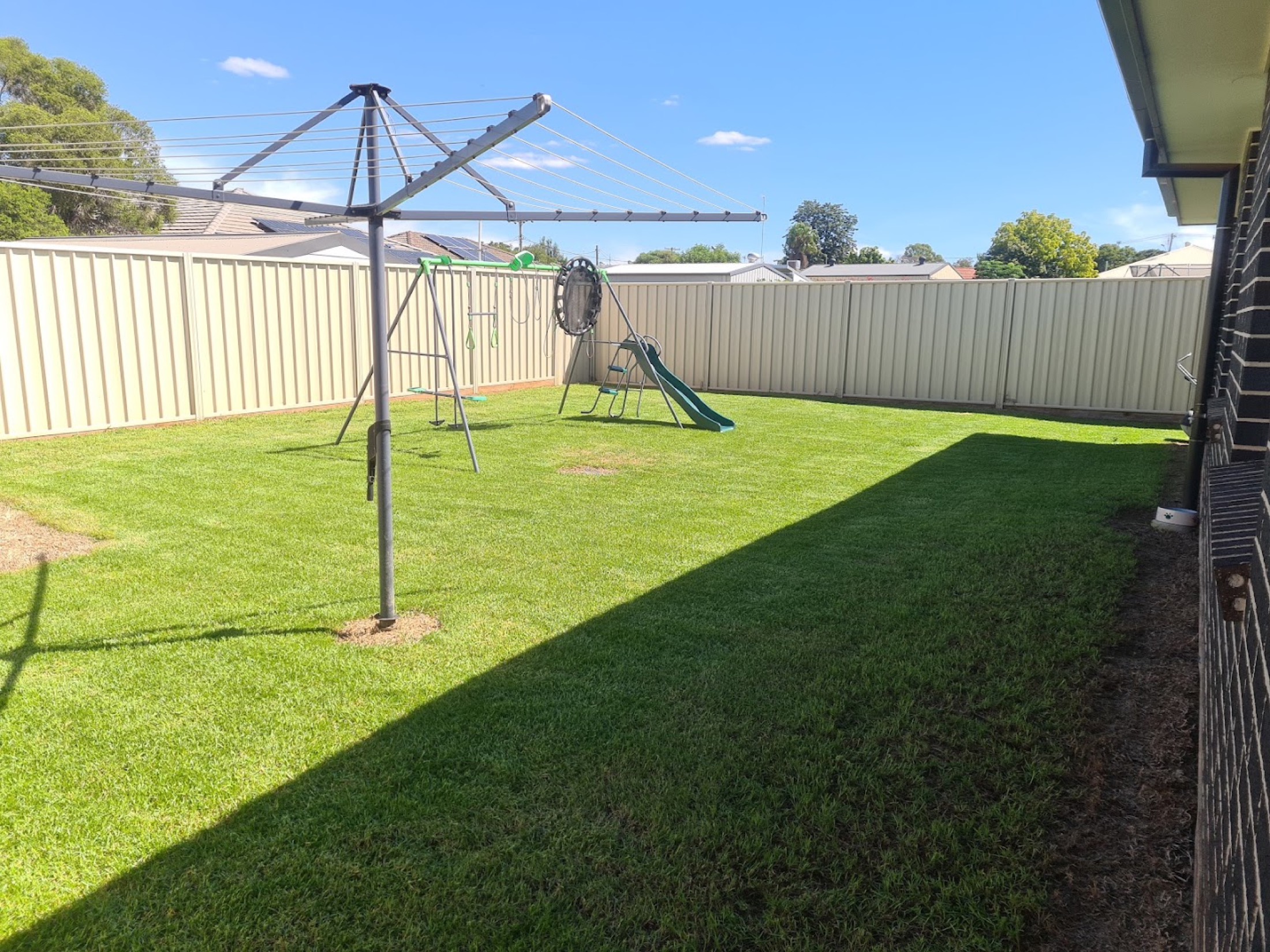 A mown backyard lawn in Dubbo with a rotary clothesline, swing set and beige Colorbond fence